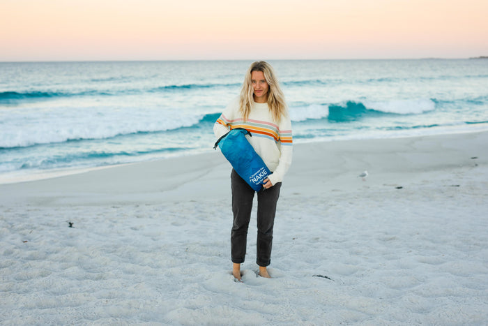 Blue sustainable down puffy blanket held by person on beach, shown with rainbow stripe sweater
