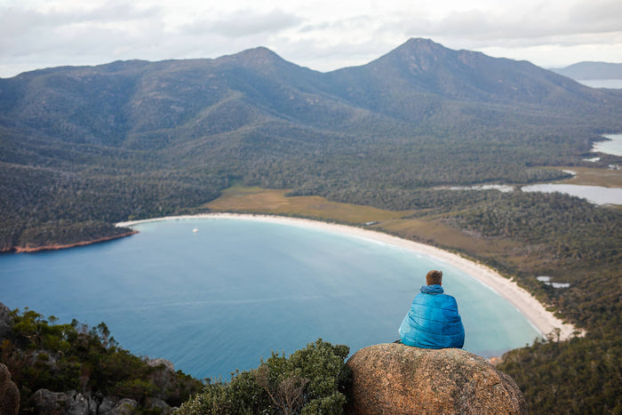 Blue quilted down puffy blanket worn by person sitting on rock overlooking coastal mountain landscape
