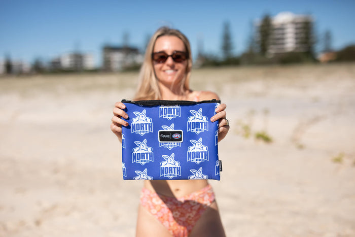 Blue North Melbourne Kangaroos AFL recycled pouch with white logo repeat pattern and black zip, held on beach.