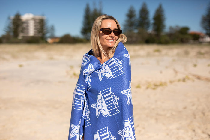 Blue North Melbourne Kangaroos AFL beach towel with white logo print, worn as wrap on sandy beach.