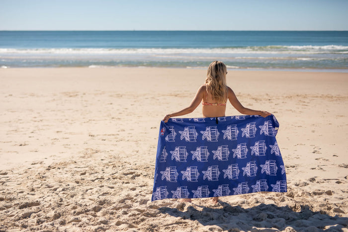 North Melbourne Kangaroos AFL beach towel in navy blue with white repeated logo pattern, draped on sandy beach.