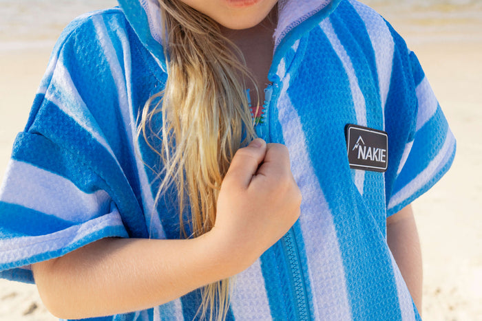 Blue and white striped kids hooded towel with Nakie brand label, shown worn by child at beach