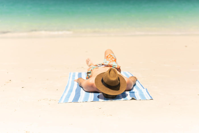 Blue and white striped recycled beach towel with person lying on sand wearing orange patterned swimwear and tan hat