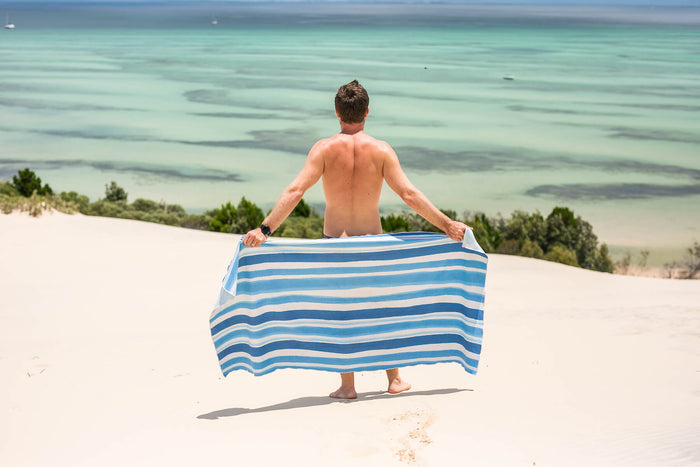 Blue and white striped recycled beach towel held by man on sandy beach overlooking turquoise ocean