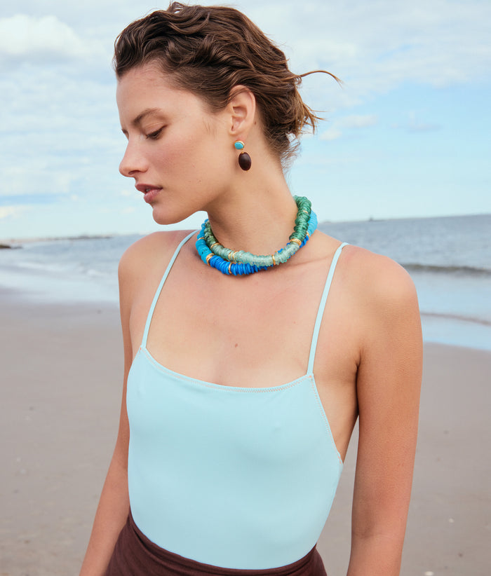 Model on beach wears blue swimsuit with Lita Necklaces in Gimlet and Azzura, and Olea Earrings