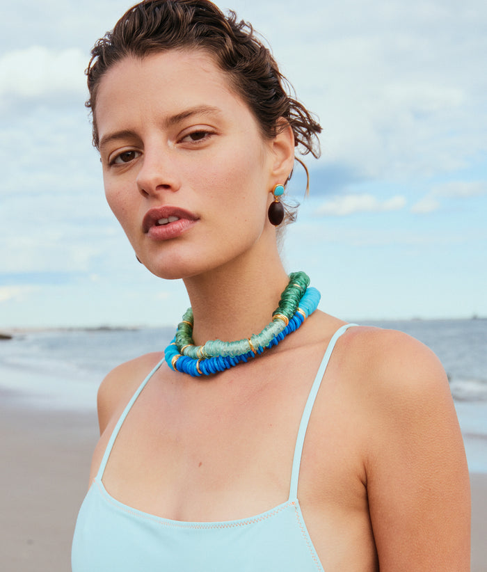 Model on beach, alt view. With blue swimsuit, Lita Necklaces in Gimlet and Azzura, and Olea Earrings