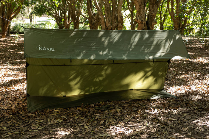 Olive ultralight tarp with NAKIE branding, pitched in forest setting with tree canopy overhead