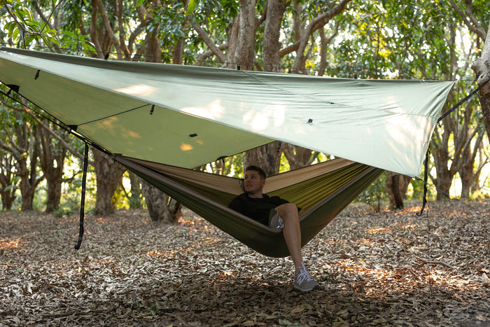 Olive ultralight tarp suspended above hammock in forest, showing no-knot setup with light-colored rain fly