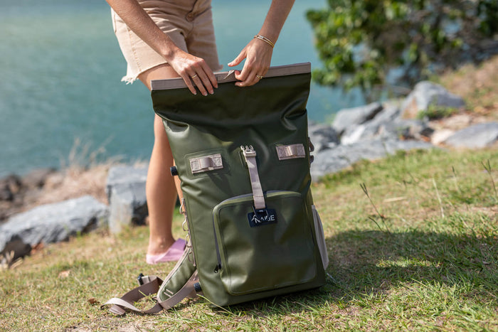 Olive green recycled cooler backpack with brown handles and straps, displayed open by the water.
