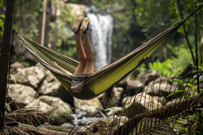 Olive green recycled hammock with tan straps suspended between trees over a forest stream with waterfall backdrop.