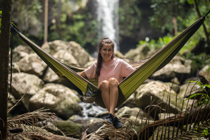Olive green recycled hammock with black straps suspended between trees above a forest stream with waterfall backdrop.
