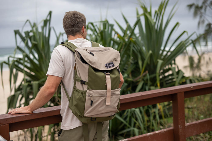 Olive green and beige recycled travel backpack with flap top and multiple compartments, worn on back by man at beach.