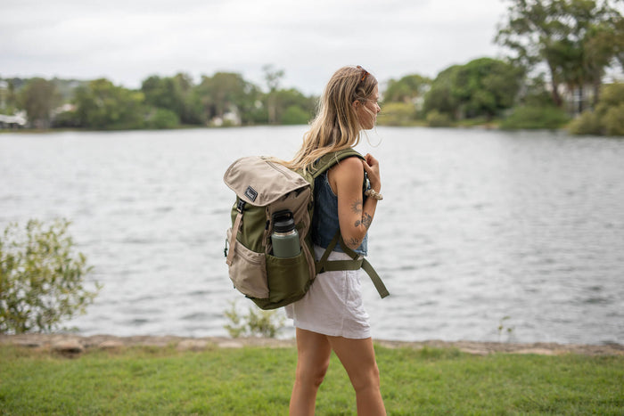 Olive green and tan recycled travel backpack with water bottle pocket, worn by woman standing at waterfront