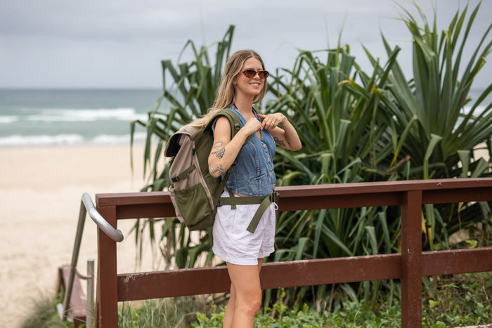Olive green recycled travel backpack worn on woman's shoulders at beach overlook with denim vest