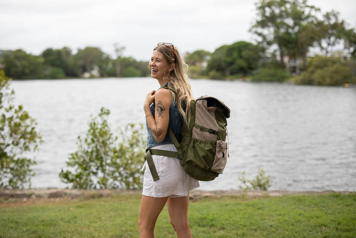Olive green recycled travel backpack with roll-top closure, worn by woman standing by water
