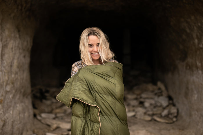 Olive green down puffy blanket wrapped around woman's shoulders, quilted design with tan trim.