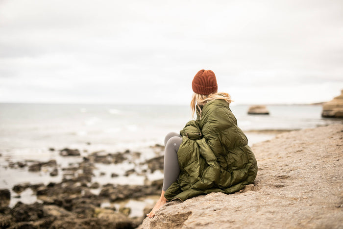 Olive green puffy down blanket draped over person sitting on beach, worn over shoulders