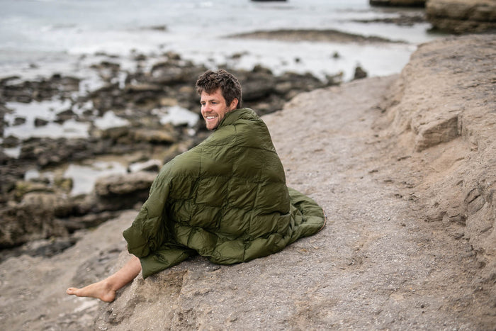 Olive green quilted down puffy blanket draped around man sitting on beach sand