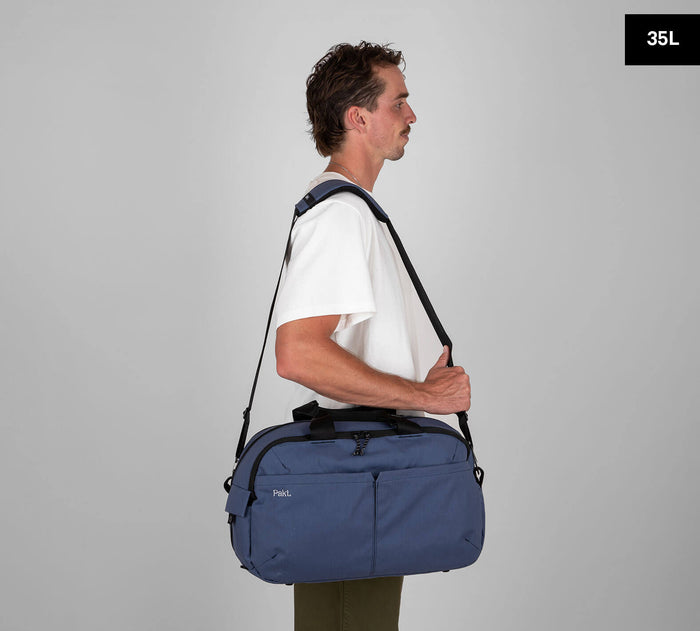 Man in white tee carrying navy-blue duffel bag over shoulder, side view; tag reads '35L'.