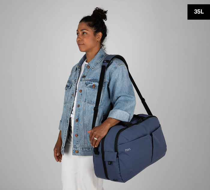 Three-quarter view of Pakt One Travel Duffel 35L in Ocean showing adjustable shoulder strap, front pockets, and carry handles on gray background