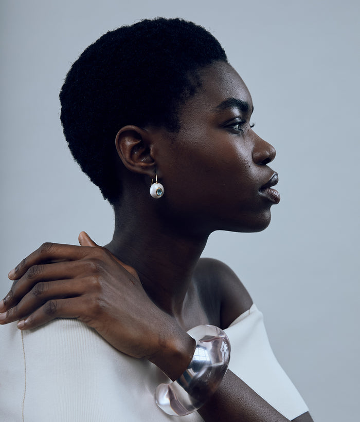 Model against grey background wearing a while top with Pearl Pablo Earrings in Blue Topaz and Arc Cuff in Clear.