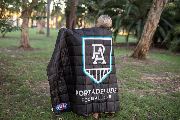 Black quilted puffy blanket with Port Adelaide Football Club logo, blue trim, and AFL badge, worn as cape outdoors.
