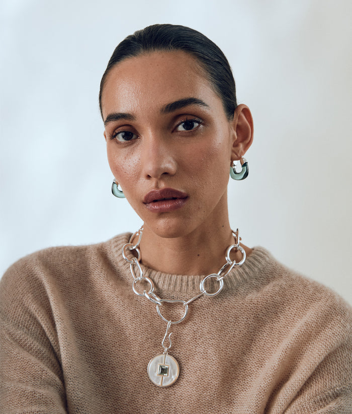 Model in tan sweater wears Porto Chain and Porto Pendant in Silver with Mini Organic Hoops in Forest & Silver.