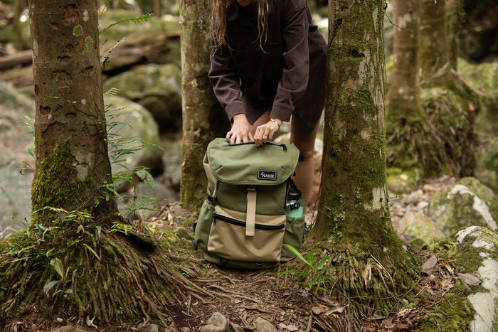 Olive-green recycled backpack with tan straps worn in forest setting between moss-covered trees