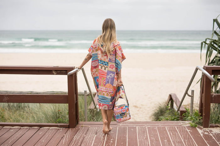 Retro Shores hooded towel in multicolor patchwork print, worn over shoulder by person on beach boardwalk.