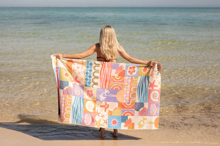 Recycled sand-free beach towel with colorful retro patchwork pattern held open by woman at the shore.