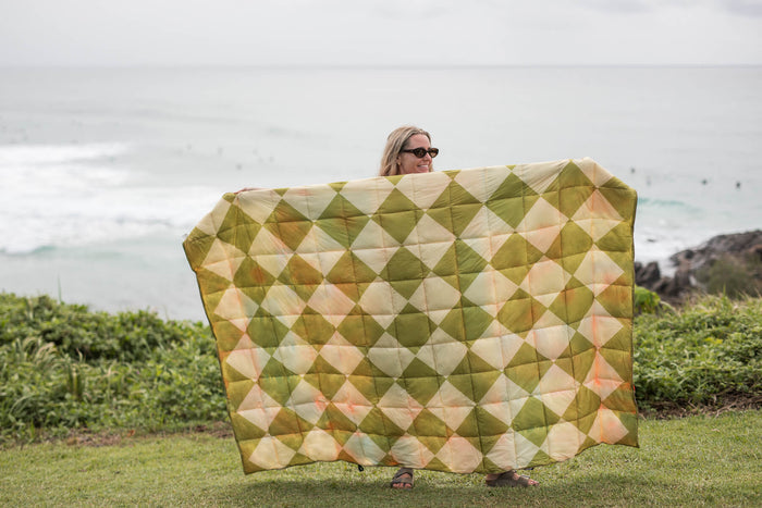 Retro Shores sustainable down puffy blanket in olive, cream, and peach diamond pattern, displayed outdoors by ocean.