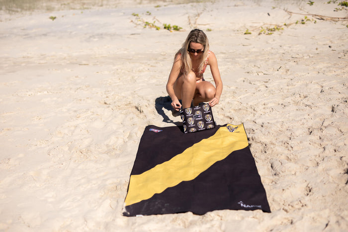 Black and gold Richmond Tigers AFL beach towel with diagonal stripe design, displayed on sand at the beach