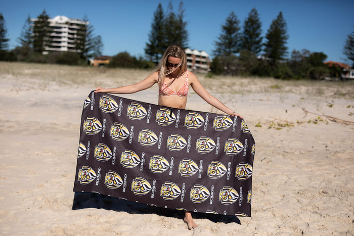 Black Richmond Tigers AFL beach towel with repeated gold and white logo pattern, held up by woman on sandy beach