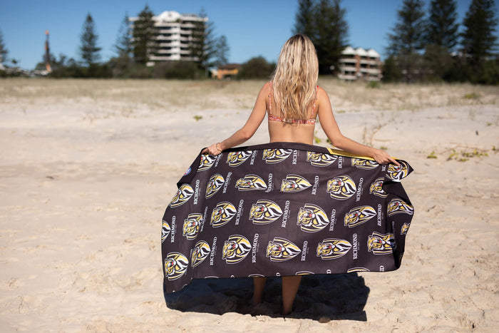 Black Richmond Tigers AFL beach towel with repeating tiger logo and text, held up on sand showing front design.