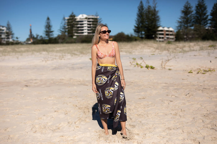 Black Richmond Tigers AFL beach towel with gold and white tiger logo print, worn as sarong-style skirt on woman at beach.