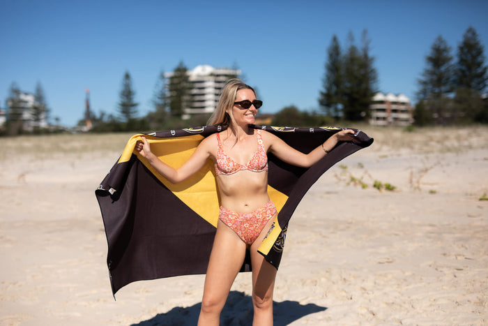 Richmond Tigers black and gold recycled beach towel with woman wearing coral floral bikini on sandy beach