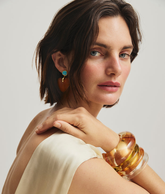 Model looks over shoulder and wears heart earrings with stack of orange and yellow resin cuffs