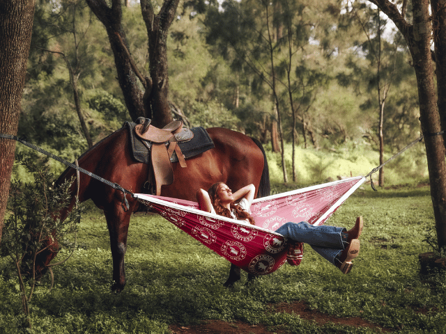 Red recycled hammock with straps suspended between trees, person relaxing underneath in forest setting.