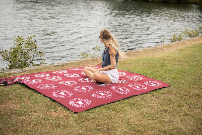 Burgundy picnic blanket with white circular Ringers Western logos spread on grass by water