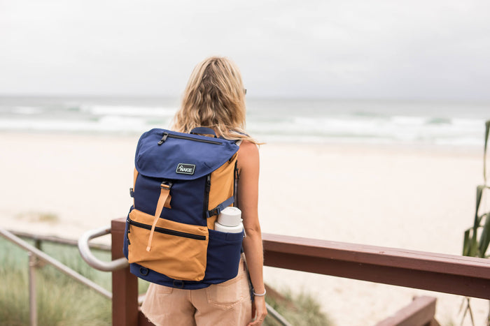 Navy and tan colorblock recycled backpack with flap top and side bottle pocket, worn by person at beach overlook