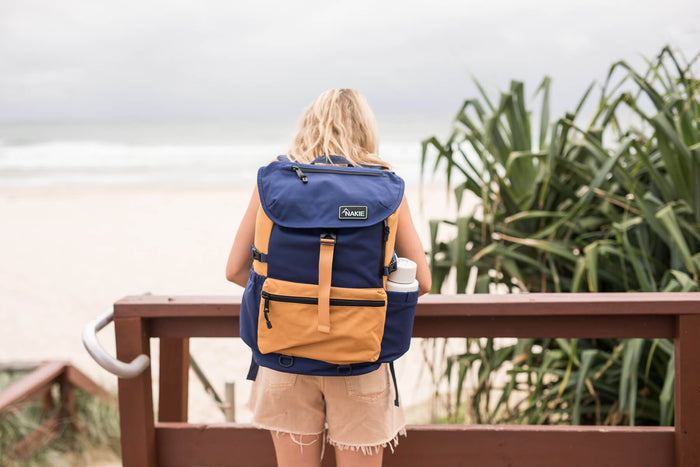 Navy and tan recycled travel backpack with flap closure, worn on back view at beach.