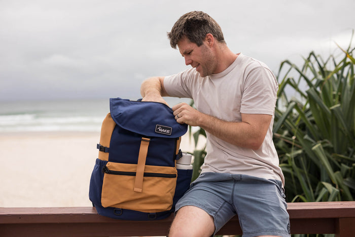 Navy and tan recycled travel backpack with flap closure and shoulder straps, displayed on man by beach
