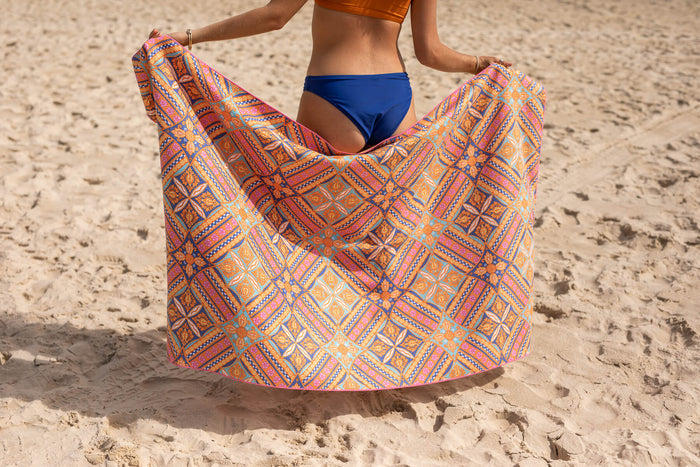 Multicolored geometric patterned beach towel in orange, pink, and blue tones, displayed on sand.