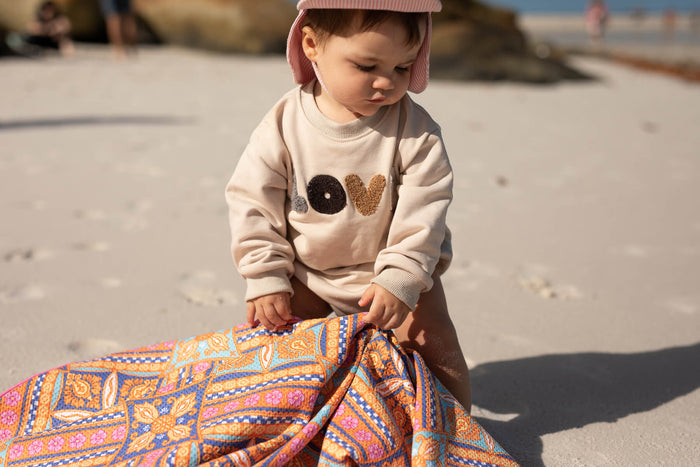 Colorful patterned beach towel in orange, blue, and pink geometric design, held by toddler wearing cream sweatshirt.