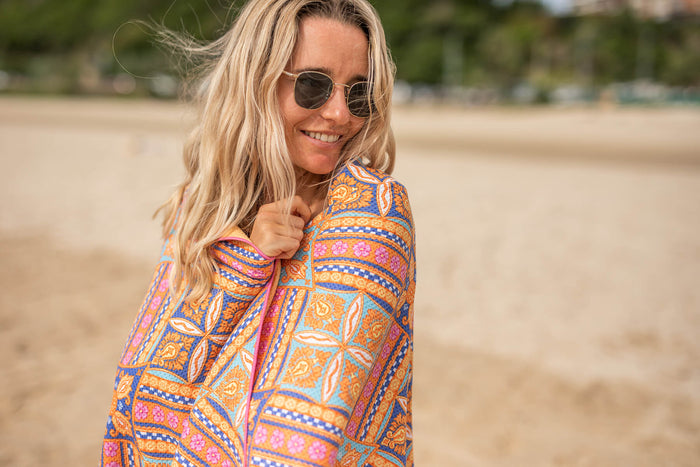 Multicoloured geometric patterned beach towel wrapped around woman at beach, gold and blue tones