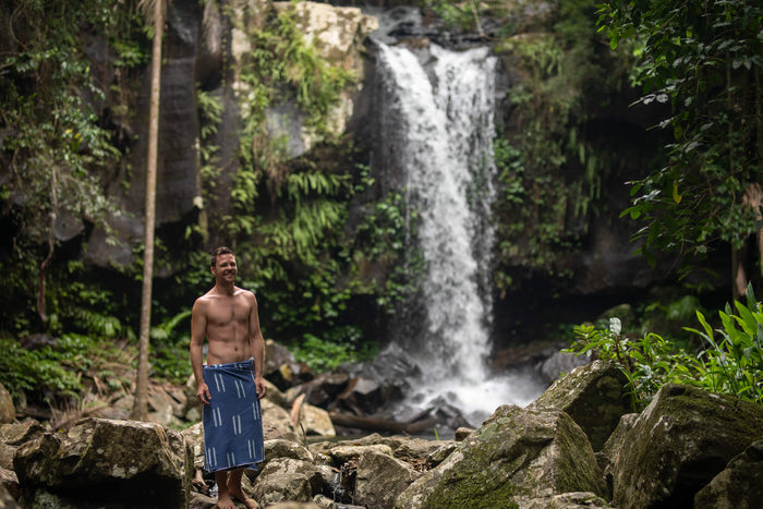 Navy beach towel with white stripe pattern, worn as wrap by man standing on rocks near waterfall