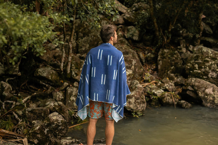 Blue recycled sand-free beach towel with white geometric stripe pattern, draped over shoulders by water's edge