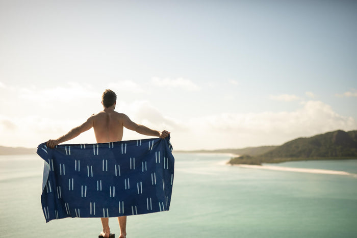 Navy beach towel with white geometric dash pattern, held open by shirtless man overlooking tropical ocean