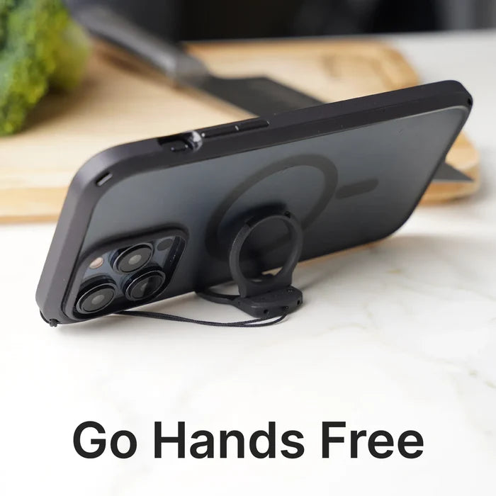 A Catalyst Case US smartphone with a Shoulder Strap & Ring Stand Bundle is propped horizontally on a white surface. A circular ring stand supports the phone, with “Go Hands Free.” in large text. A cutting board and broccoli appear in the background.