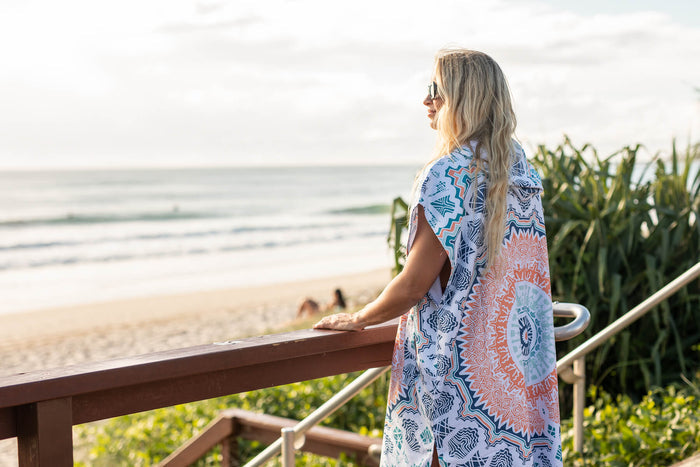 Hooded towel with blue and coral geometric print, worn over shoulder at beach overlooking ocean
